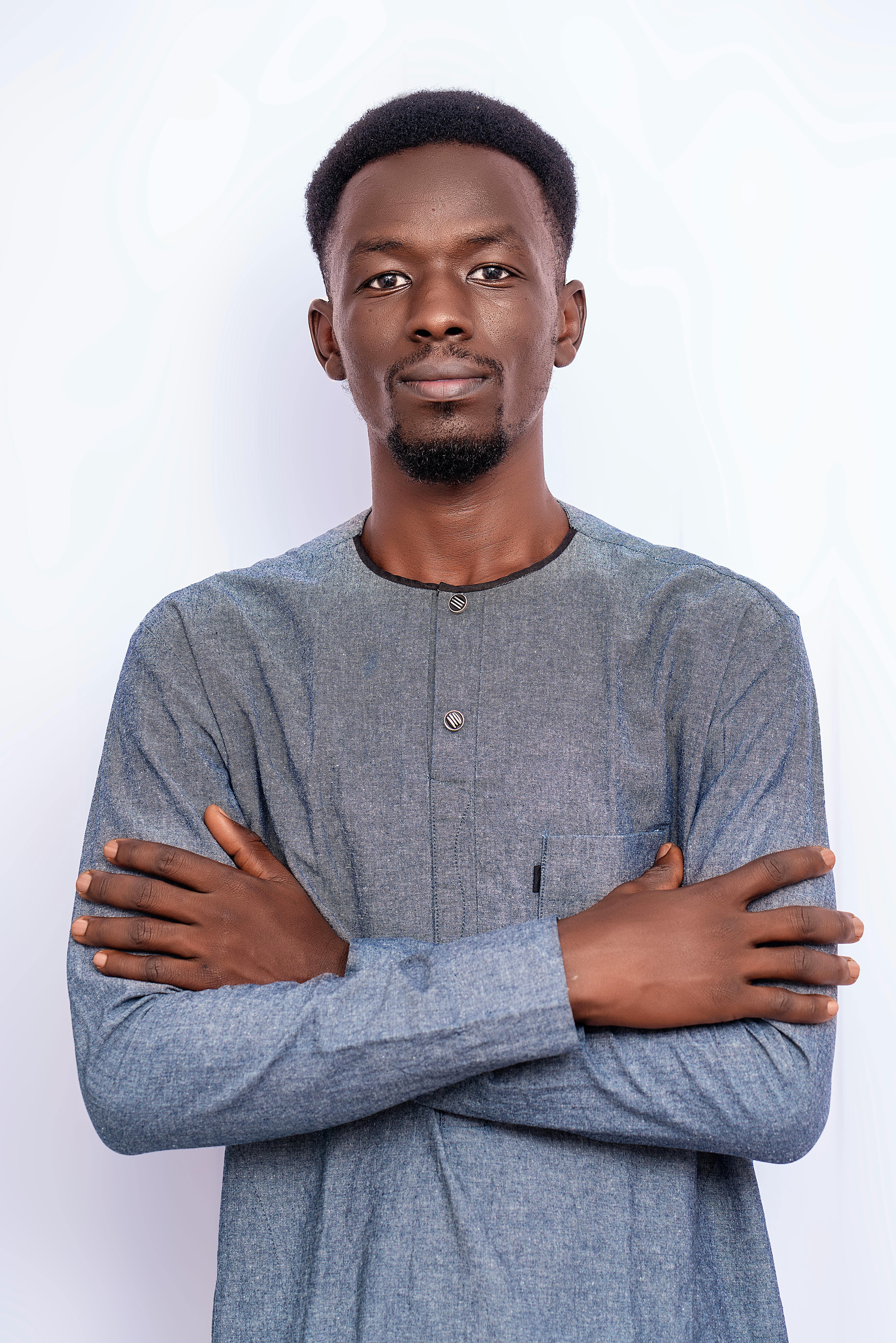 A young man with crossed arms wearing a traditional grey shirt poses confidently against a white background.