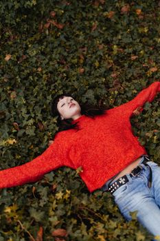 Young woman lying on leafy ground in a red sweater, embracing nature's tranquility.
