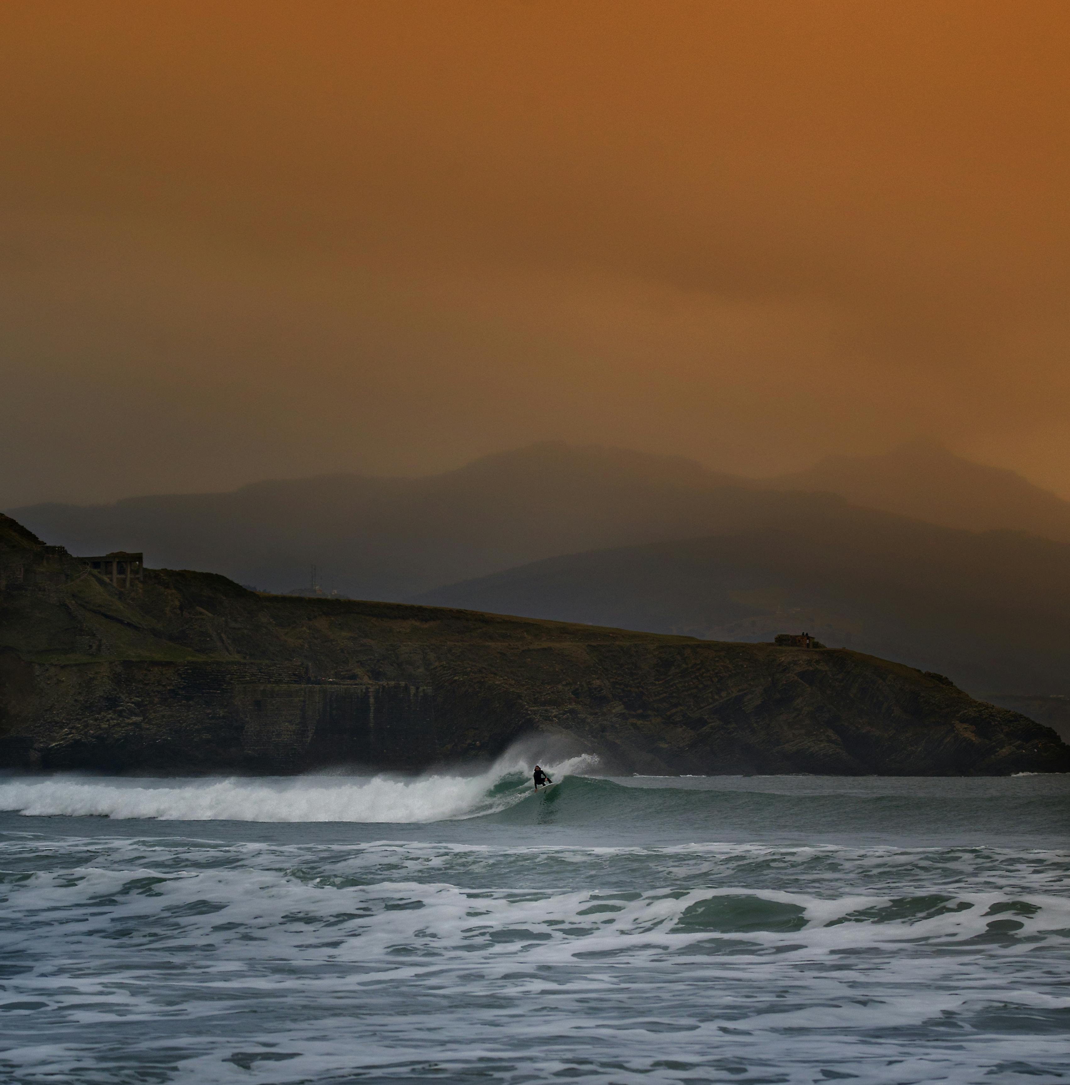 一名孤独的冲浪者迎着绚丽的橙色天空，在崎岖的海岸线上冲浪