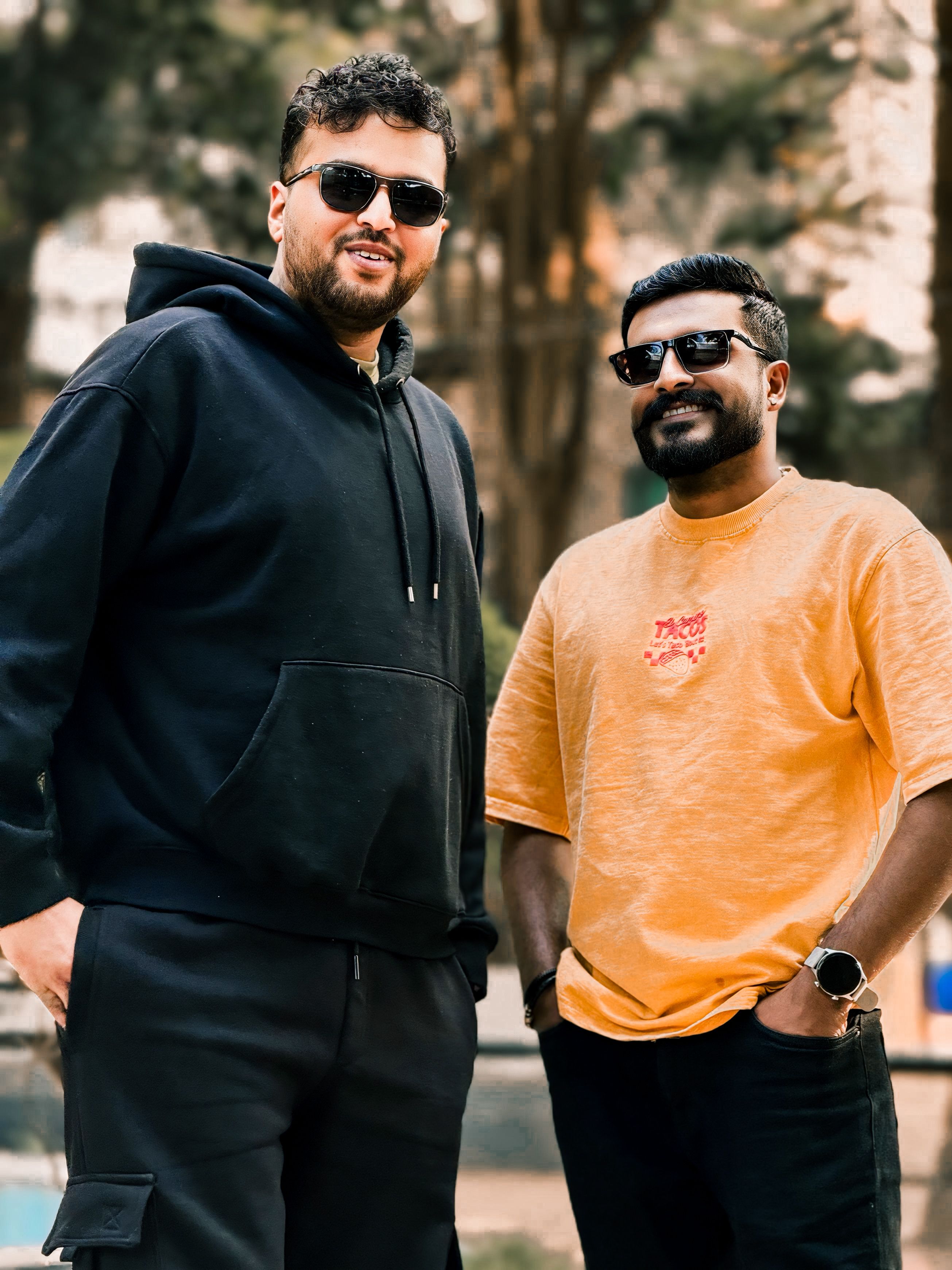 Two adult men smiling and posing outdoors wearing sunglasses in a casual setting.