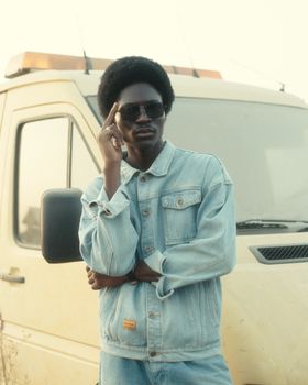 A fashionable man poses confidently in denim, standing by a van under daylight.