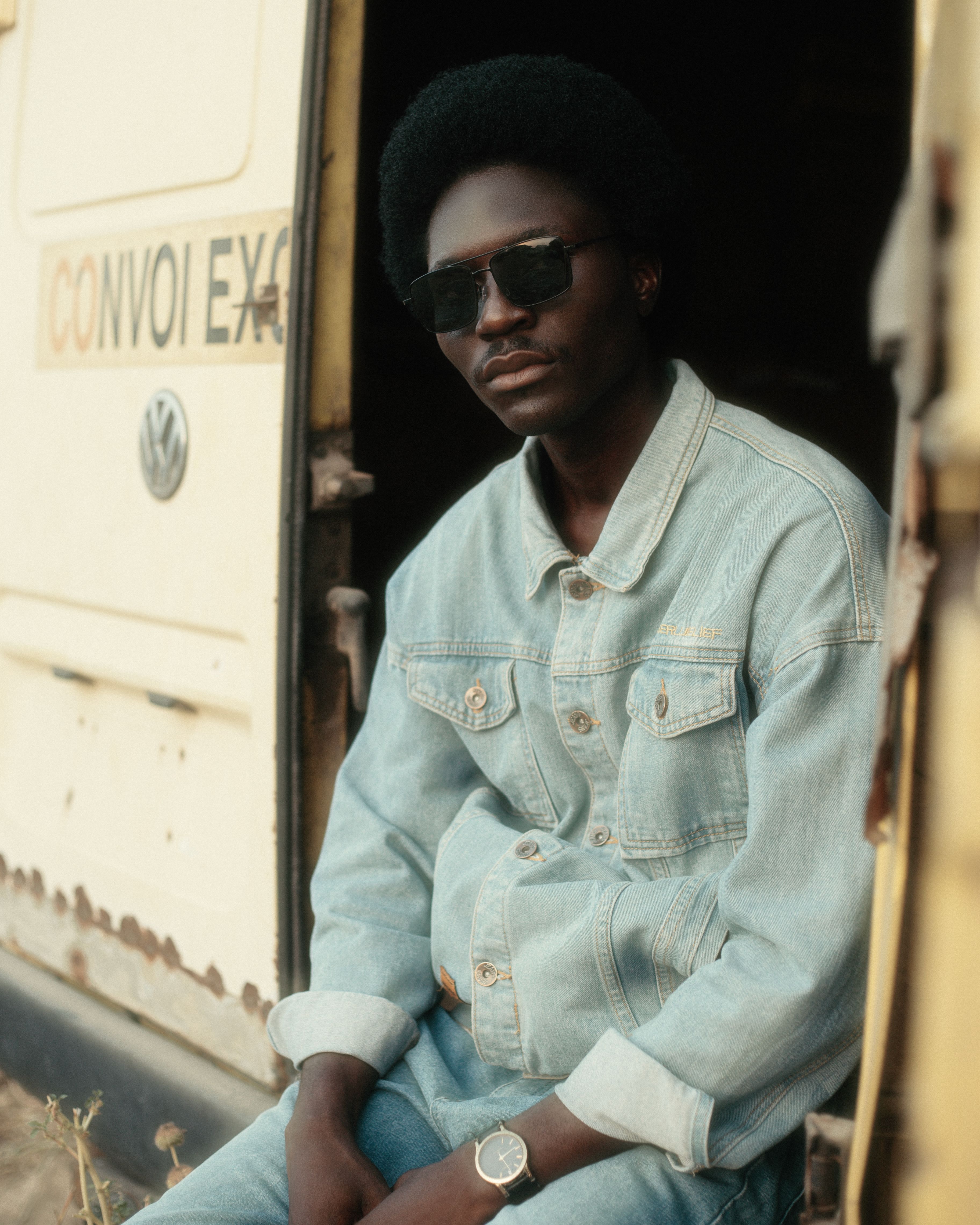 Casual portrait of a man wearing sunglasses and denim jacket seated by a vintage van.