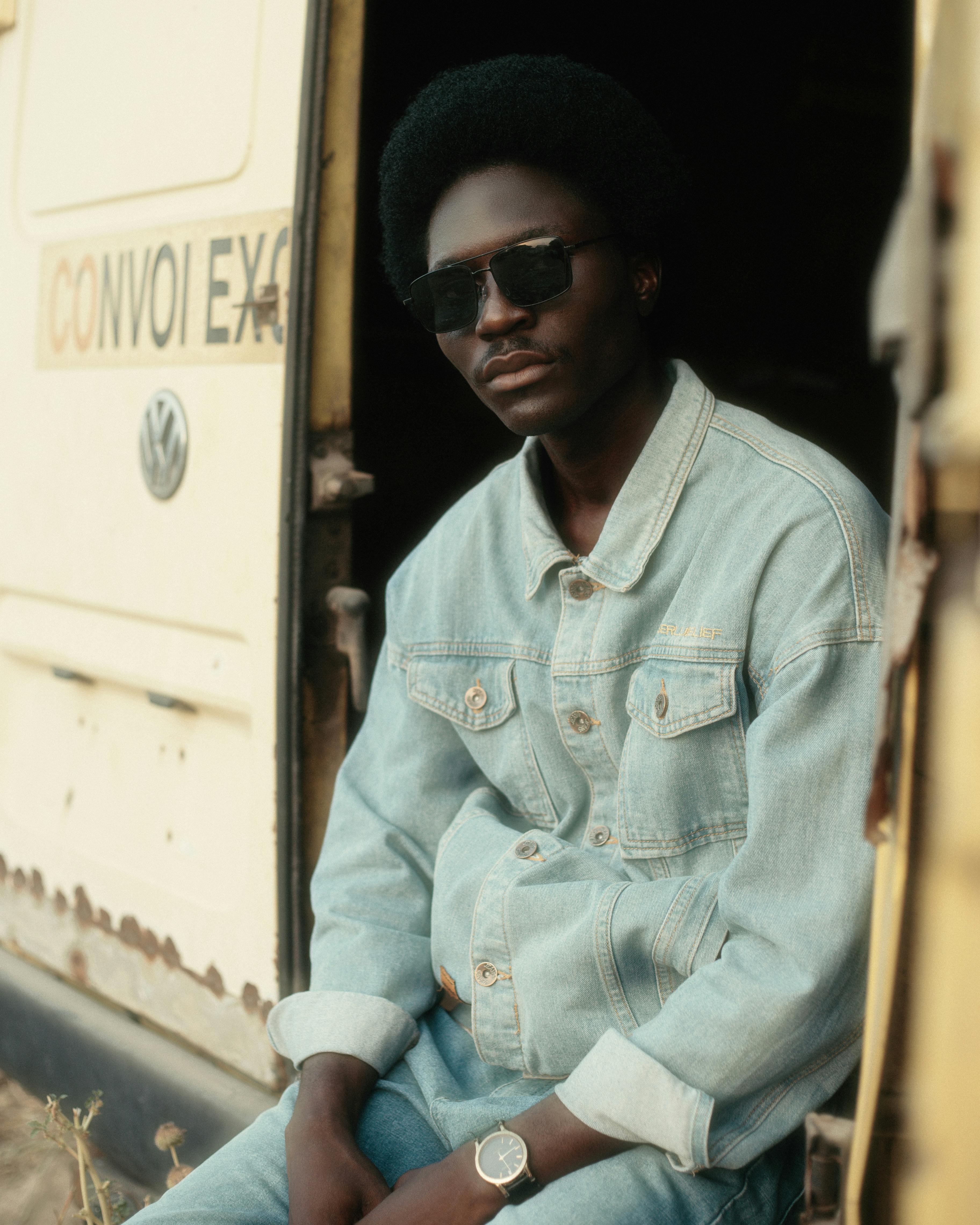 Gratis Retrato casual de un hombre con gafas de sol y chaqueta vaquera sentado junto a una furgoneta vintage. Foto de stock