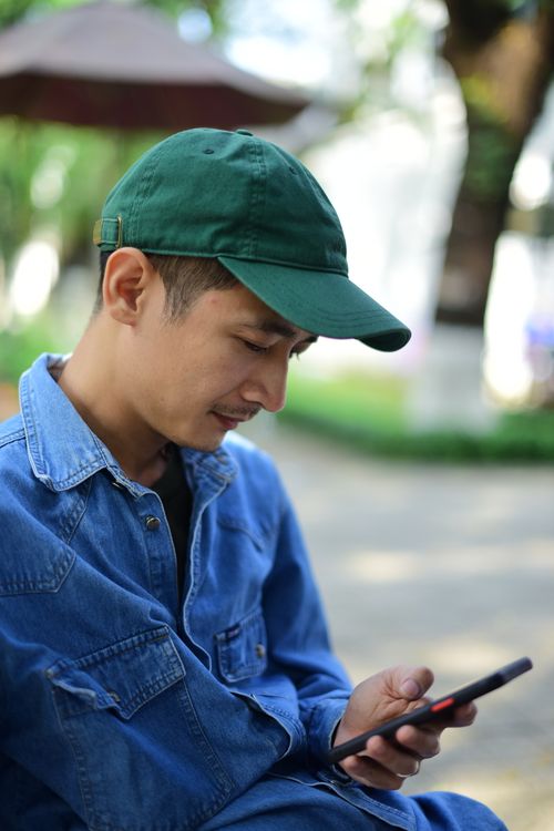 Free Young man in denim and cap texting on smartphone while sitting outdoors. Stock Photo