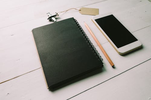 Плоское фото черной спиральной книги возле коричневого карандаша и белого смартфона