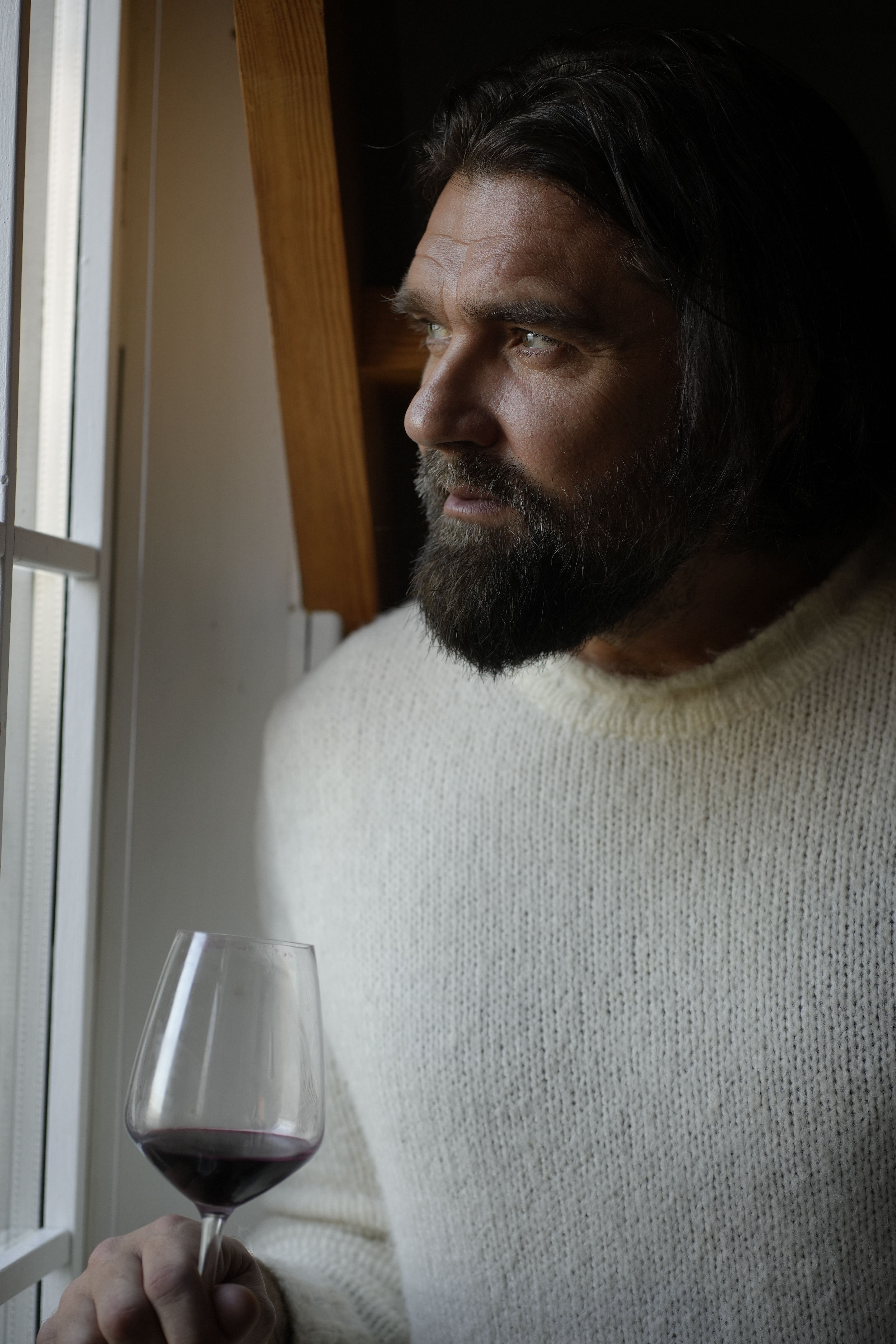Thoughtful man in sweater with wine, gazing out window indoors.