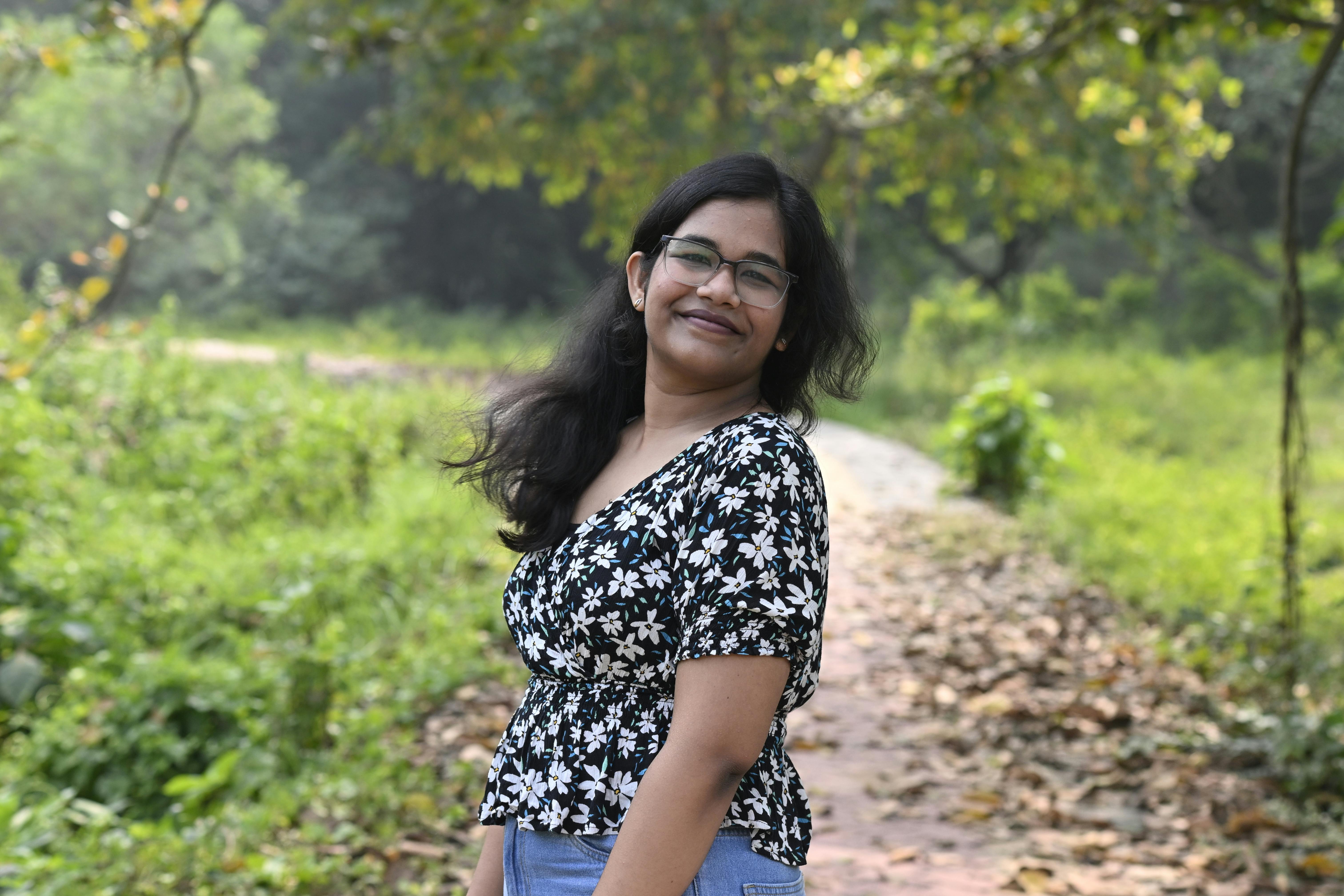 A woman smiling outdoors on a leafy path amid lush greenery.