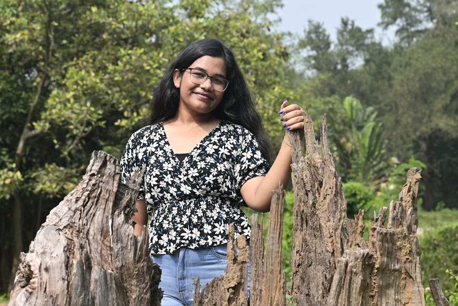 A young woman with glasses stands smiling in a sunny, natural forest setting, surrounded by tree trunks.
