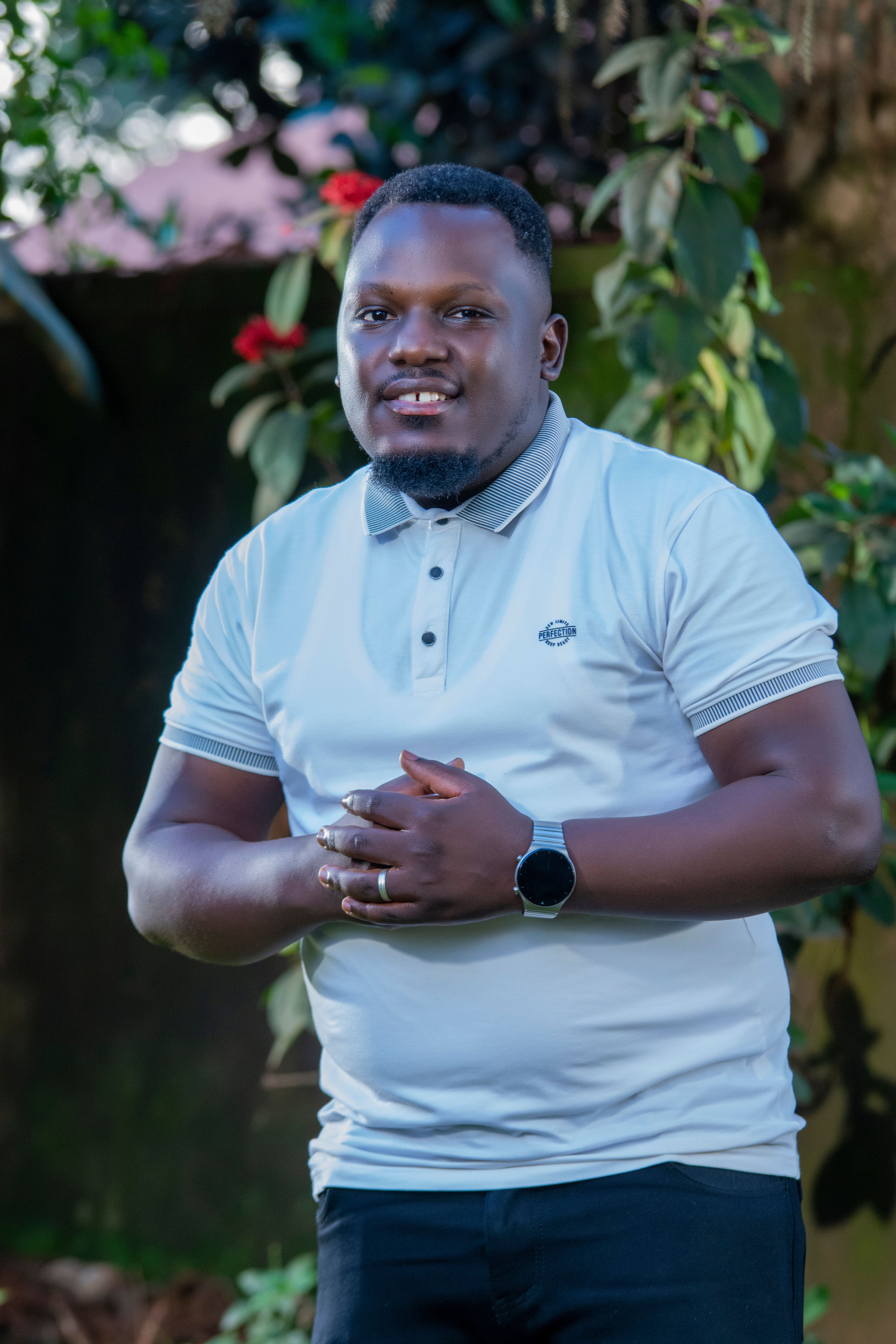 Smiling man in casual attire posing in a lush outdoor setting with greenery.