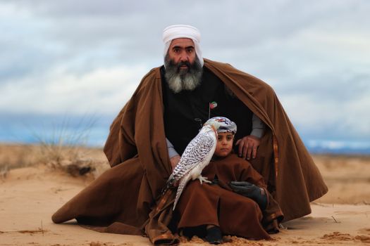 A father and child sitting with a falcon in the Algerian desert, showcasing traditional attire.