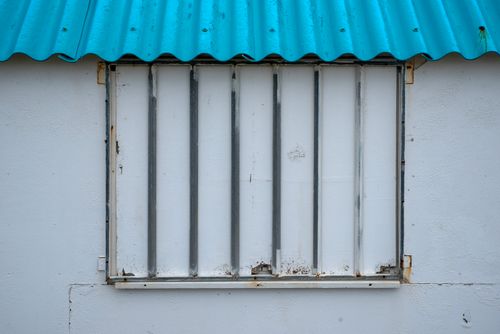 Free A weathered metal shutter on a textured white wall with a blue roof. Stock Photo