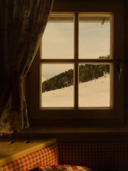 Warm interior view through a window overlooking a winter ski resort with snow-covered hills.