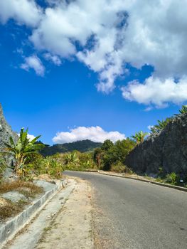 A winding road cuts through rocky hills with banana plants, under a vibrant blue sky.