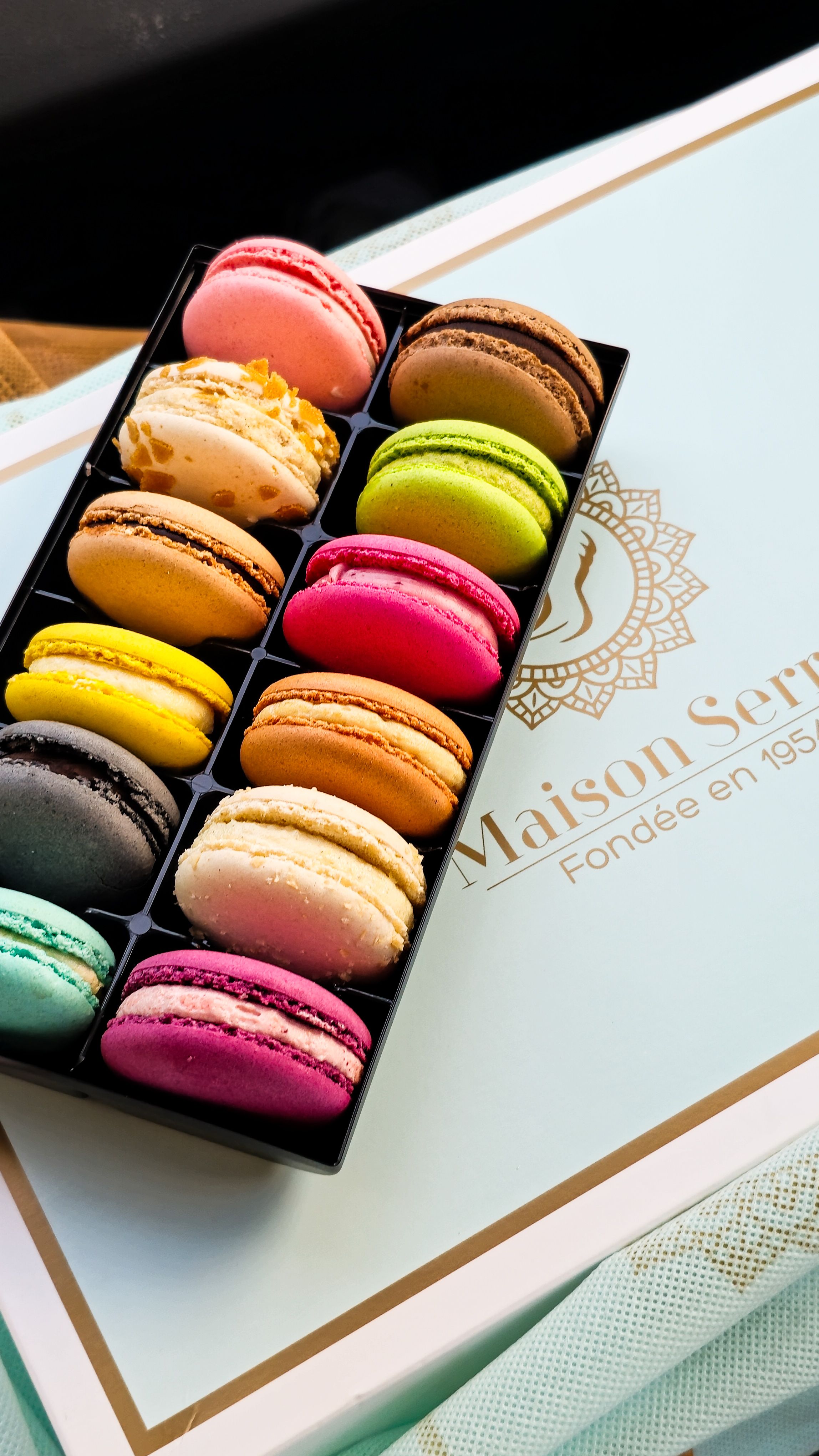Assorted colorful macarons displayed in an elegant black tray on a Maison Serre box.