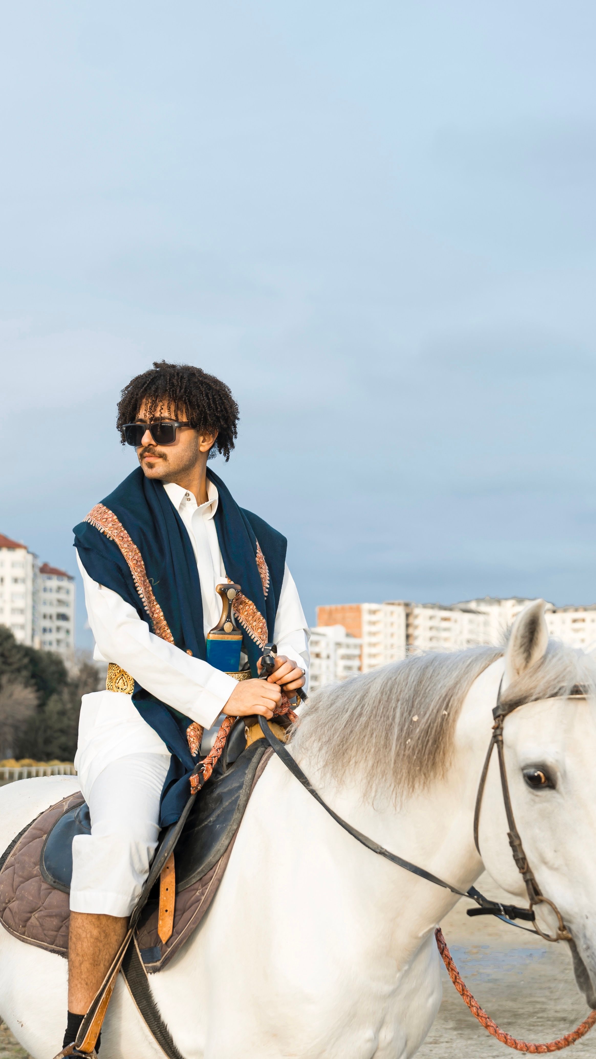 A stylish man with sunglasses rides a white horse in urban setting.