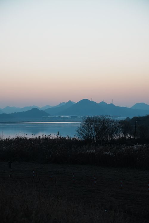 Free A tranquil scene capturing the calm water at sunset with mountains silhouetted against the pastel sky. Stock Photo