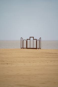 A minimalist metal sculpture stands on a sandy beach with the ocean in the background.