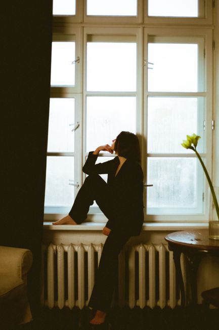 Woman in black outfit gazing out a window in a warmly lit room, evoking thoughtfulness and tranquility.