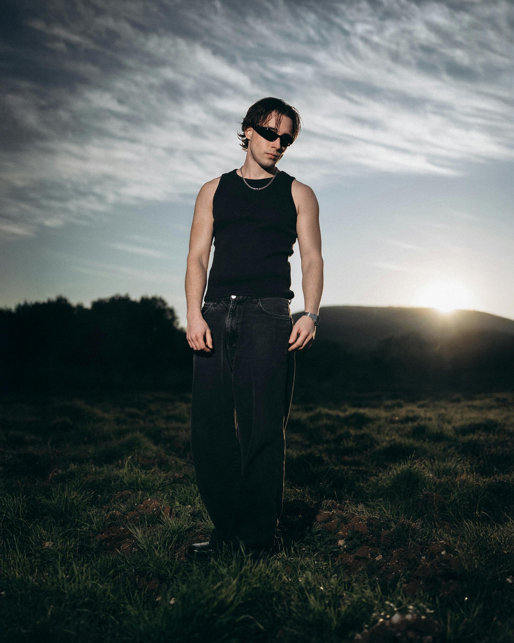 Kostenlos Ein Mann mit Sonnenbrille steht während des Sonnenuntergangs auf einem Feld; er trägt ein schwarzes Oberteil und Jeans. Stock-Foto