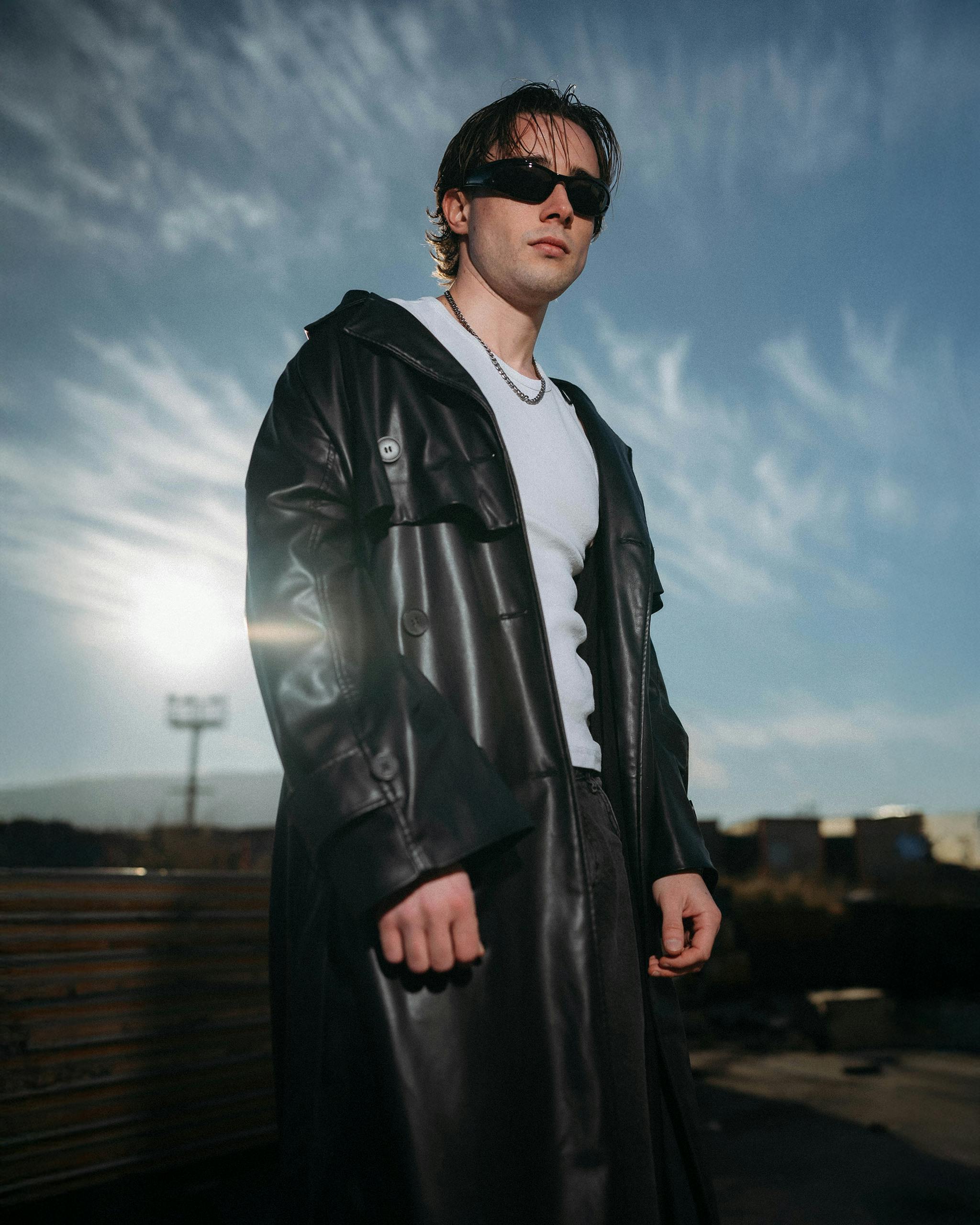 Kostenlos Selbstbewusster junger Mann mit Sonnenbrille und Ledermantel steht unter einem dramatischen Himmel. Stock-Foto