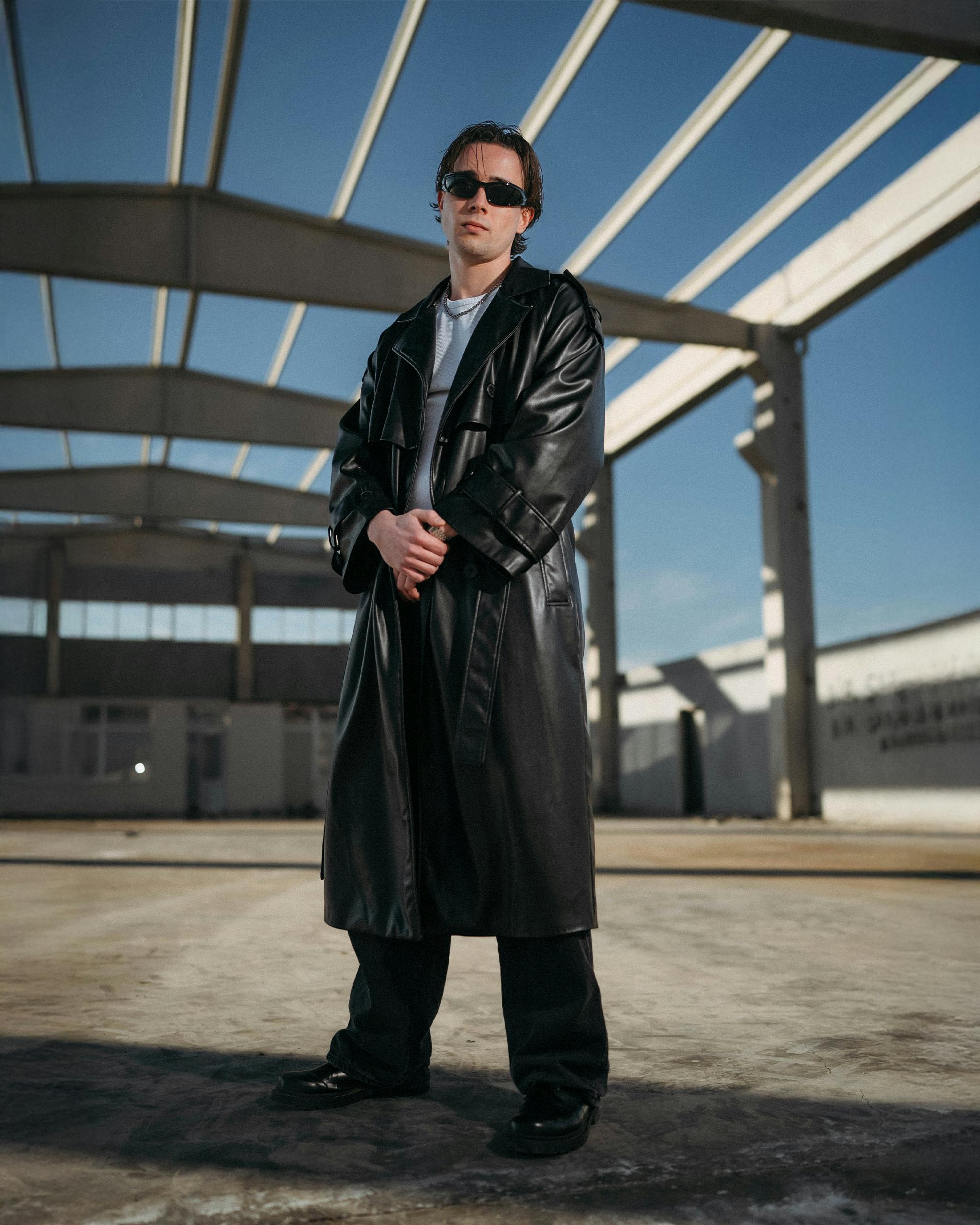 Kostenlos Ein Mann mit Sonnenbrille und Ledermantel steht selbstbewusst unter dem Gerüst eines Gebäudes. Stock-Foto