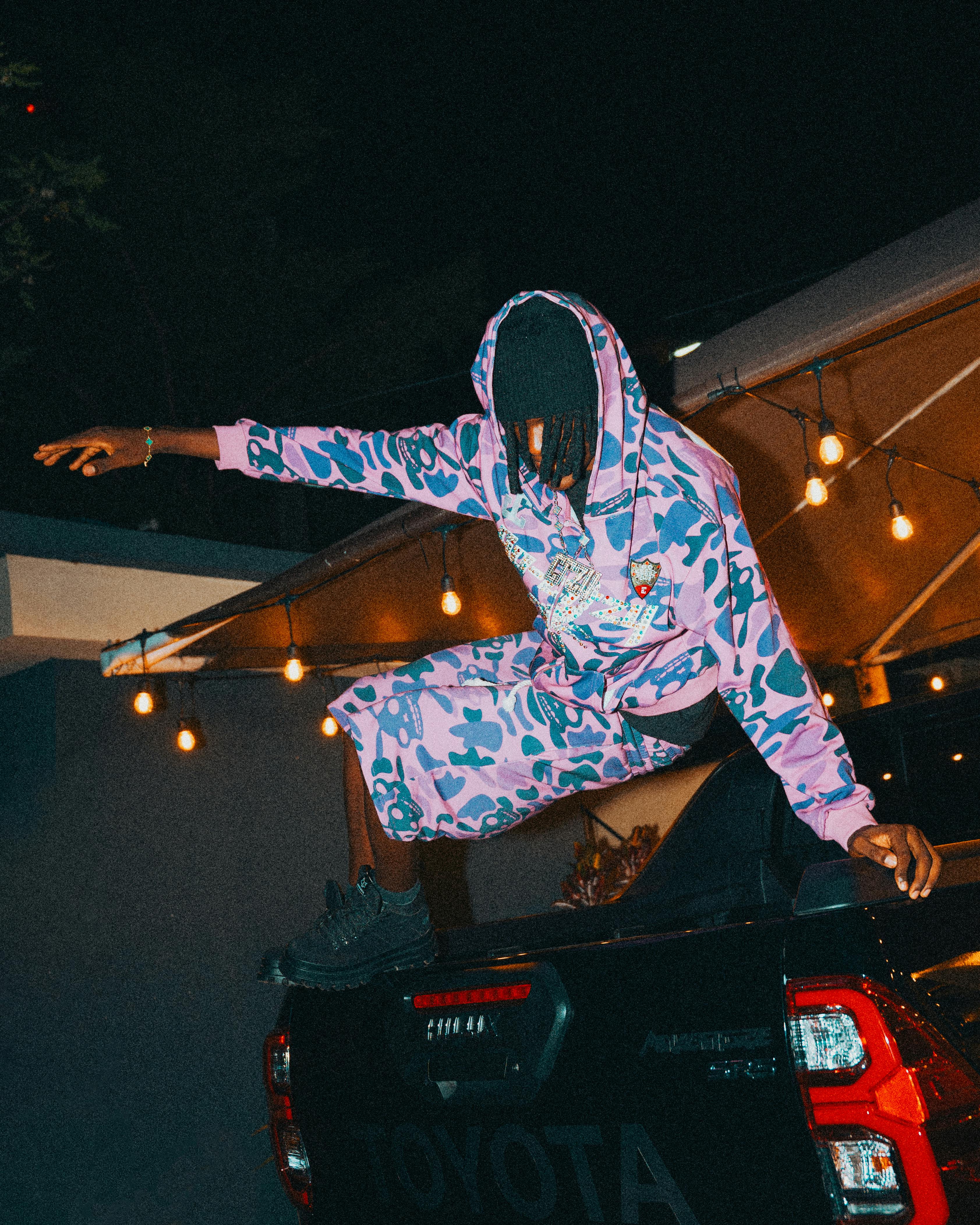 Gratis Stilig person i kamuflasjeantrekk på bil, omgitt av lyslenker om natten. Arkivbilde