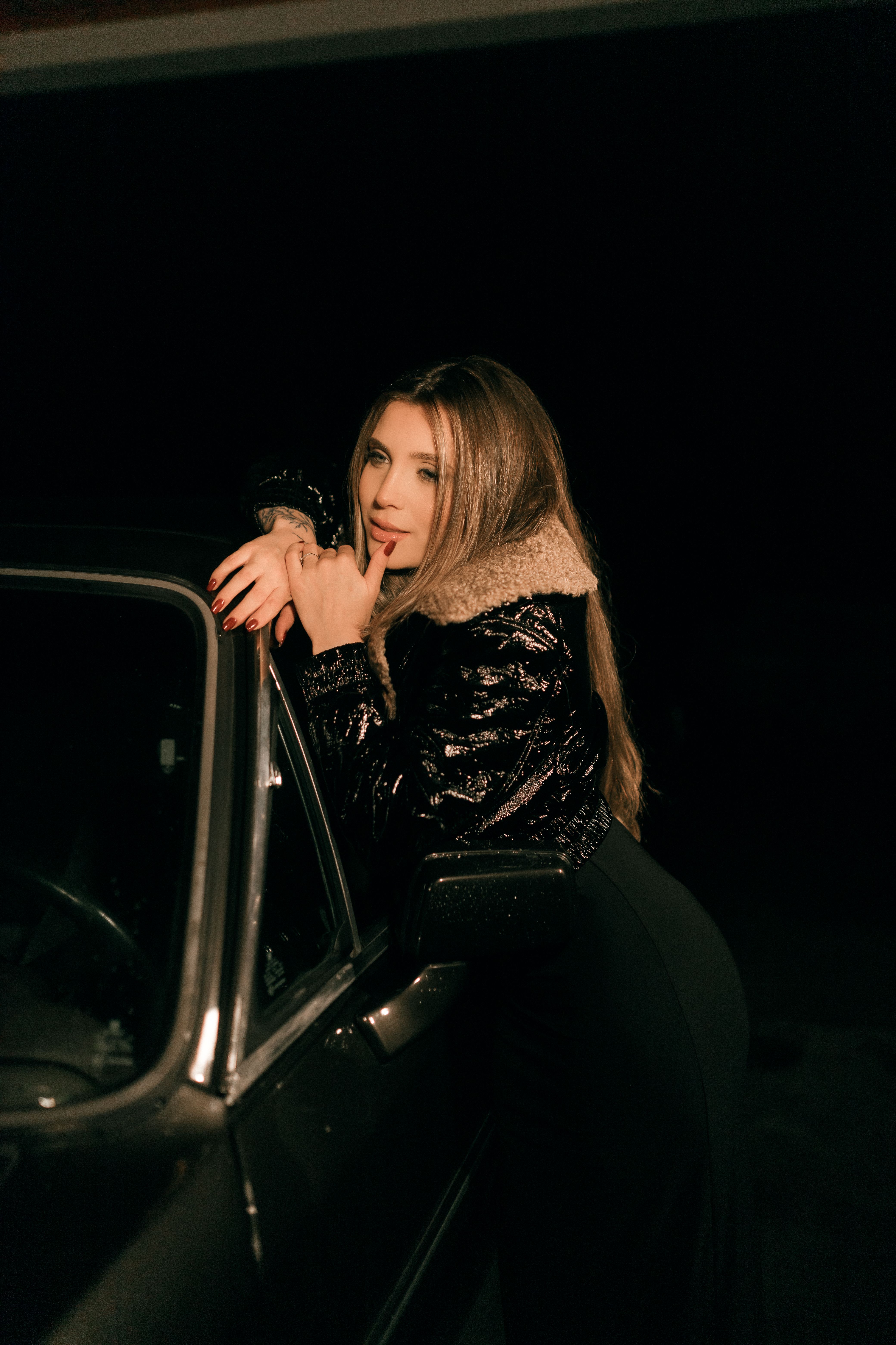 Stylish woman with long hair poses on a car against a dark nighttime backdrop.