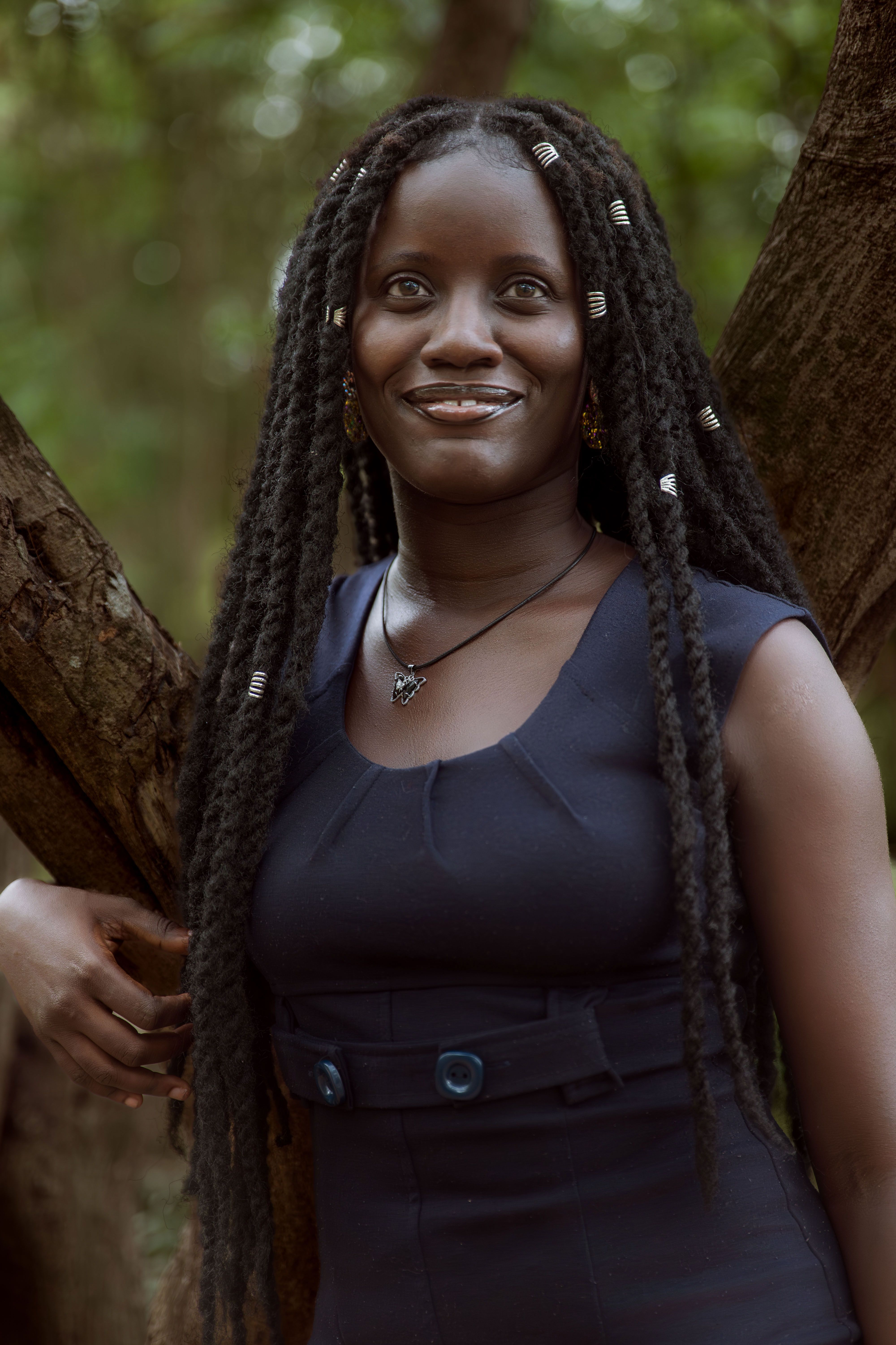 A joyful woman with long braids and a navy dress smiles while standing against a tree outdoors.