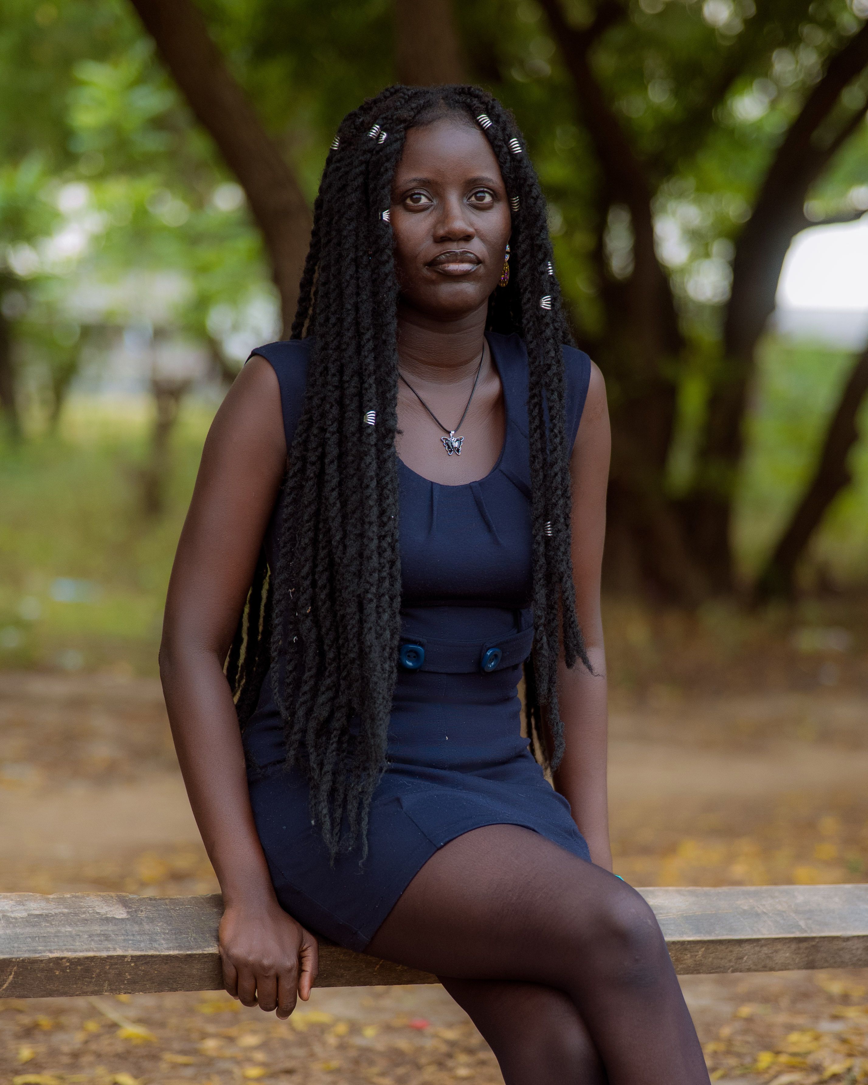 A young woman with braided hair in a blue dress sitting on a bench in a serene park environment.