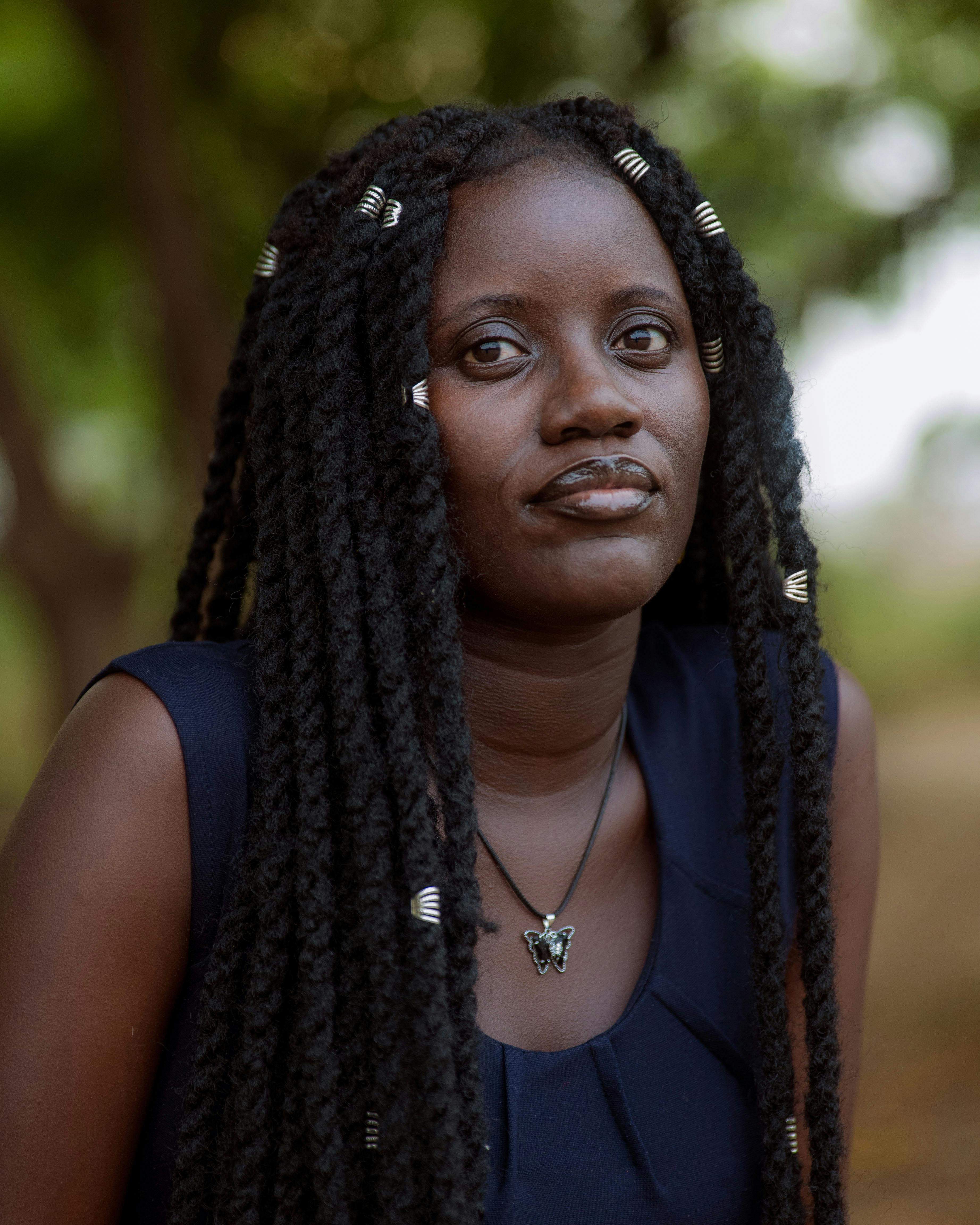 A thoughtful portrait of a woman with braided hair, captured outdoors with lush greenery in the background.