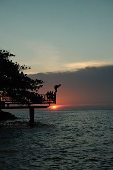 Dramatic silhouette of people diving into the ocean from a pier at sunset.