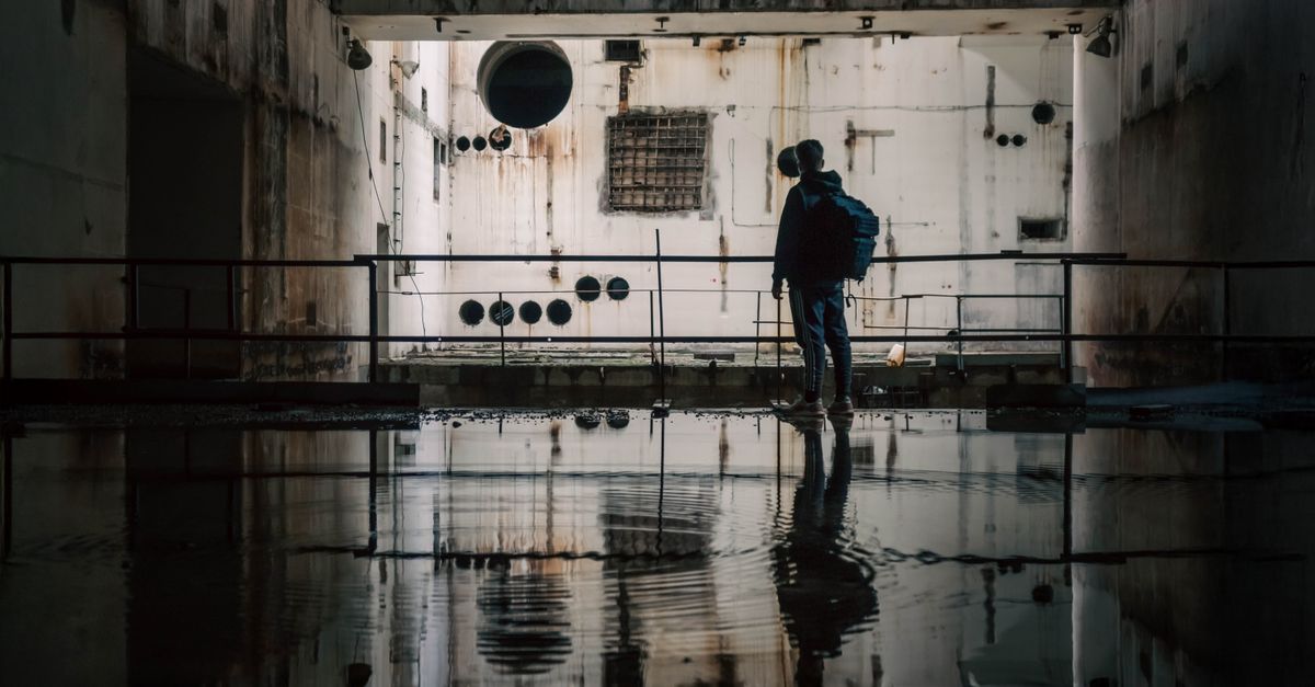 Uma pessoa explorando uma instalação industrial escura e abandonada com reflexos na água.
