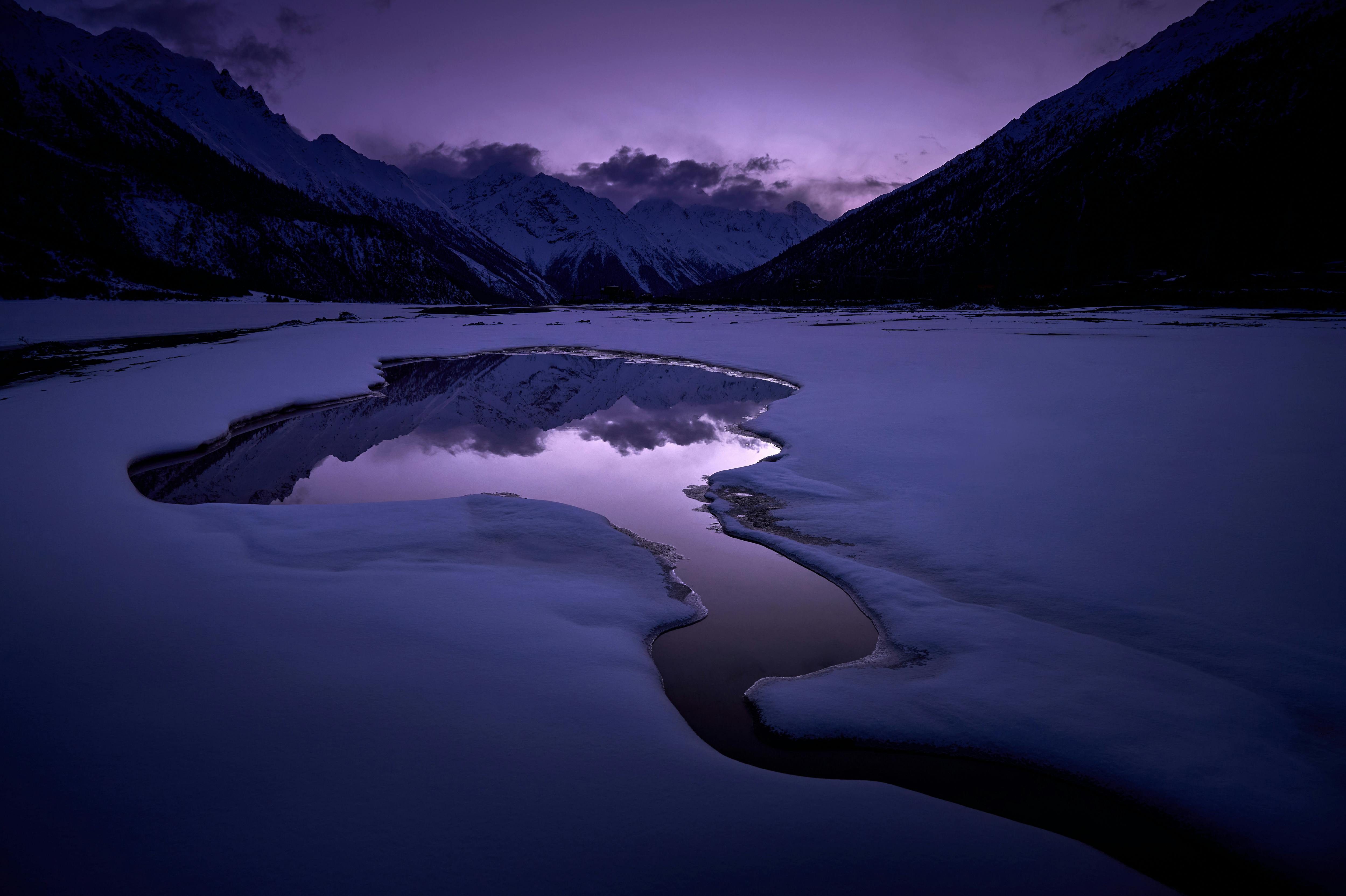 Serene twilight view of snow-covered mountains reflected in a tranquil lake, under a purple sky.