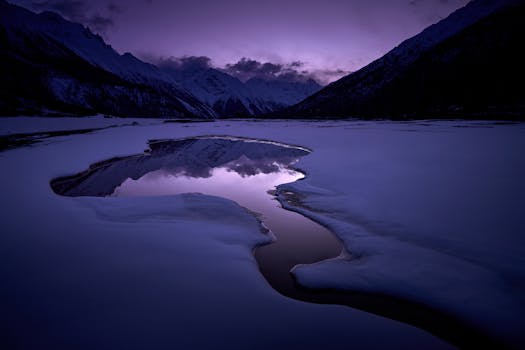 Serene twilight view of snow-covered mountains reflected in a tranquil lake, under a purple sky.