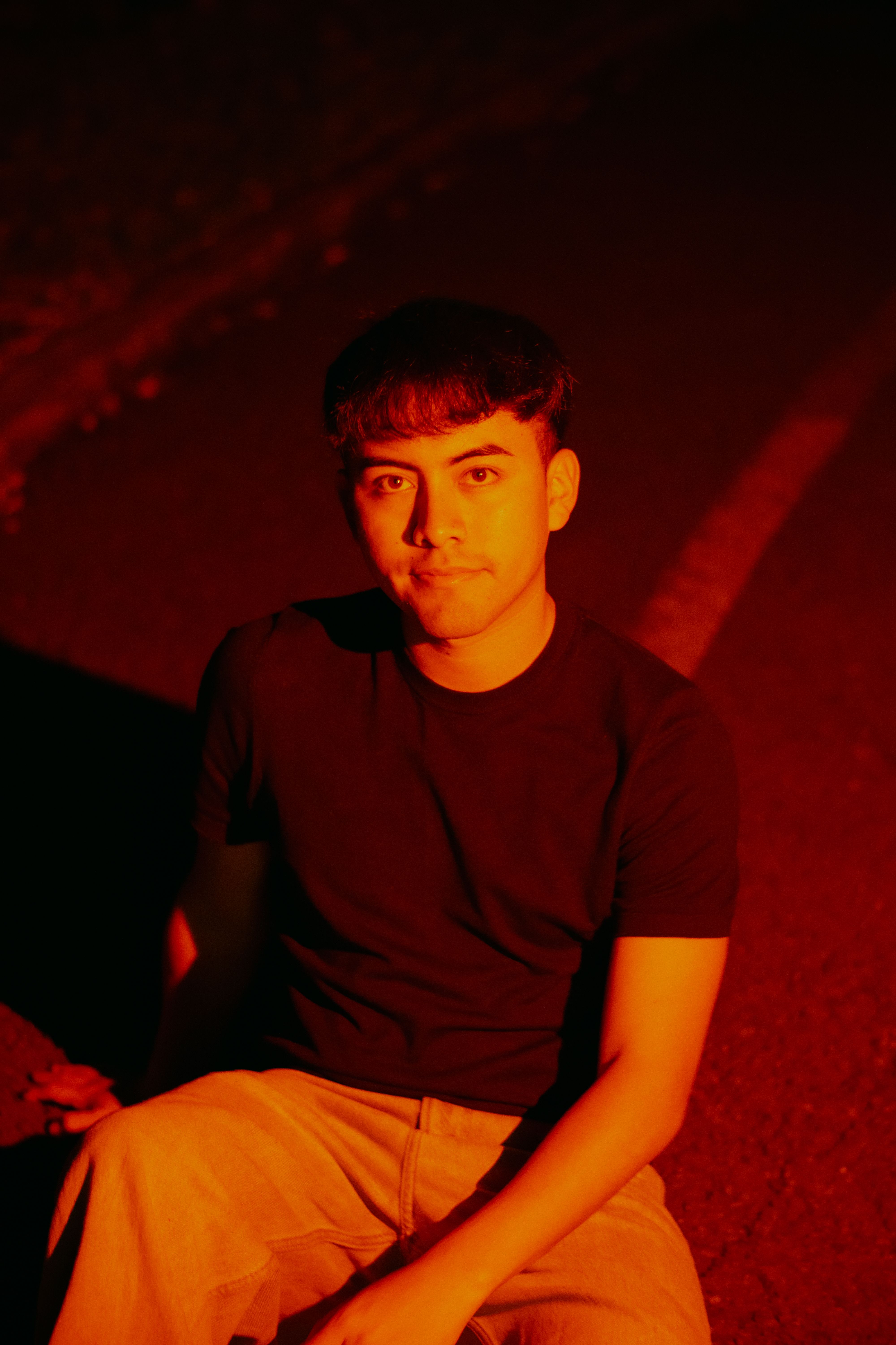 A young Hispanic man sits outdoors at night, illuminated by dramatic red lighting.