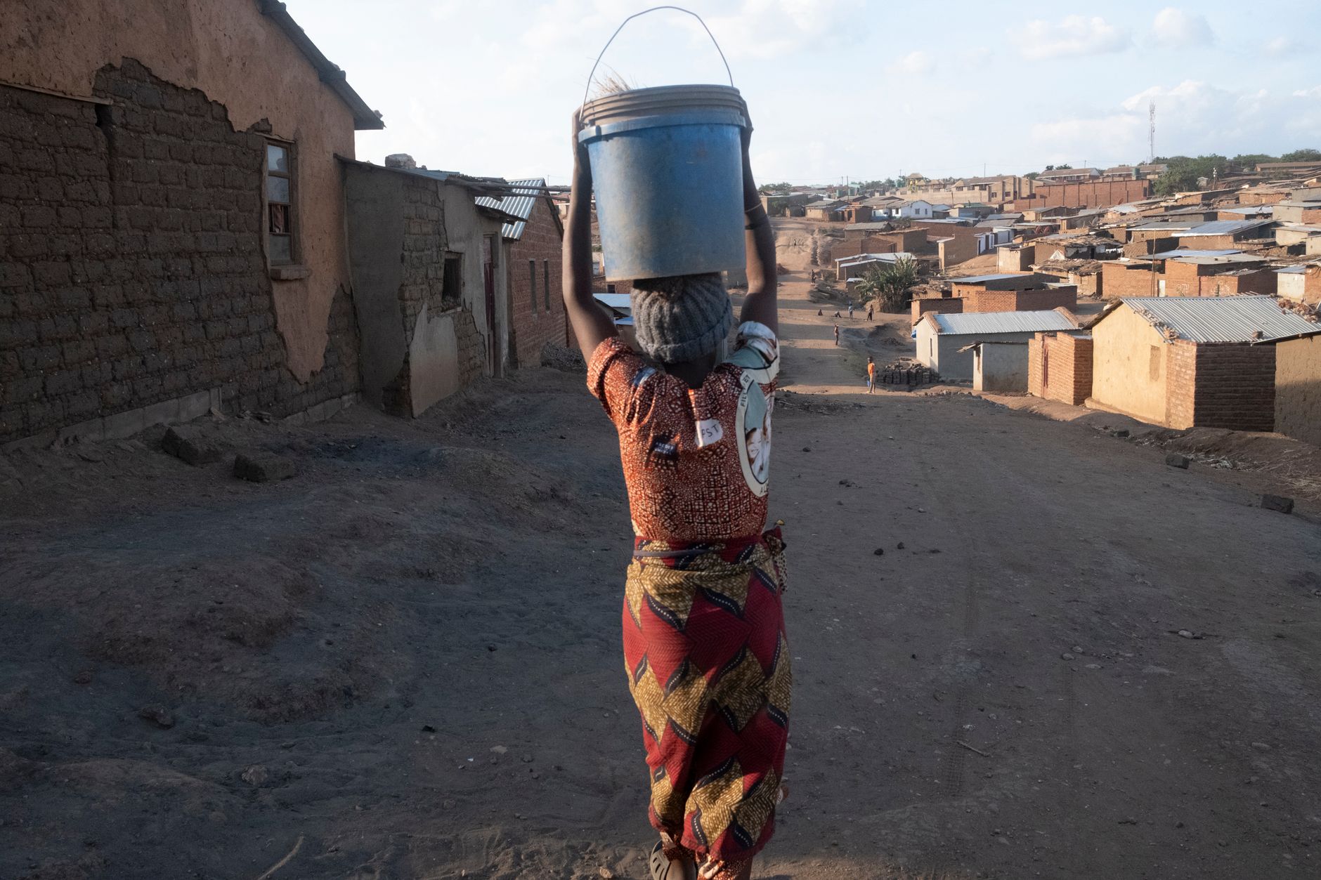 A woman balances a water bucket on her head in a rural village setting.