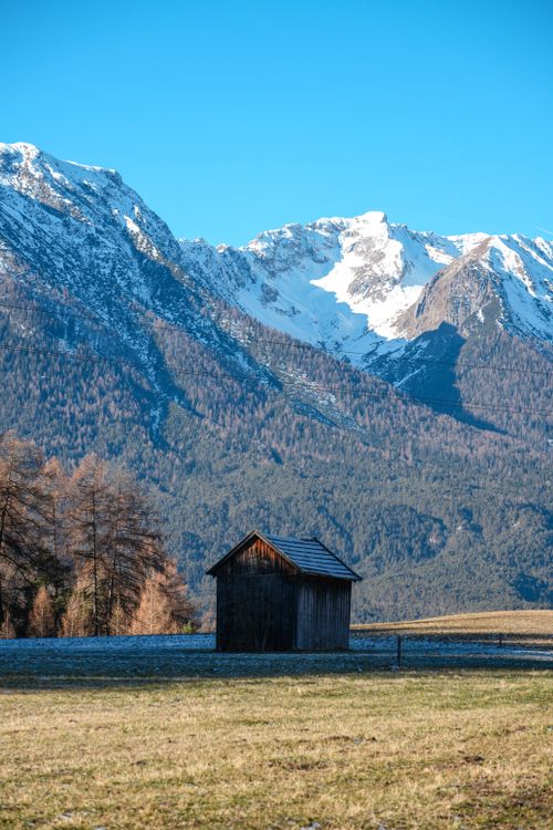 Free A solitary wooden cabin surrounded by majestic snow-capped mountains under a clear blue sky. Stock Photo