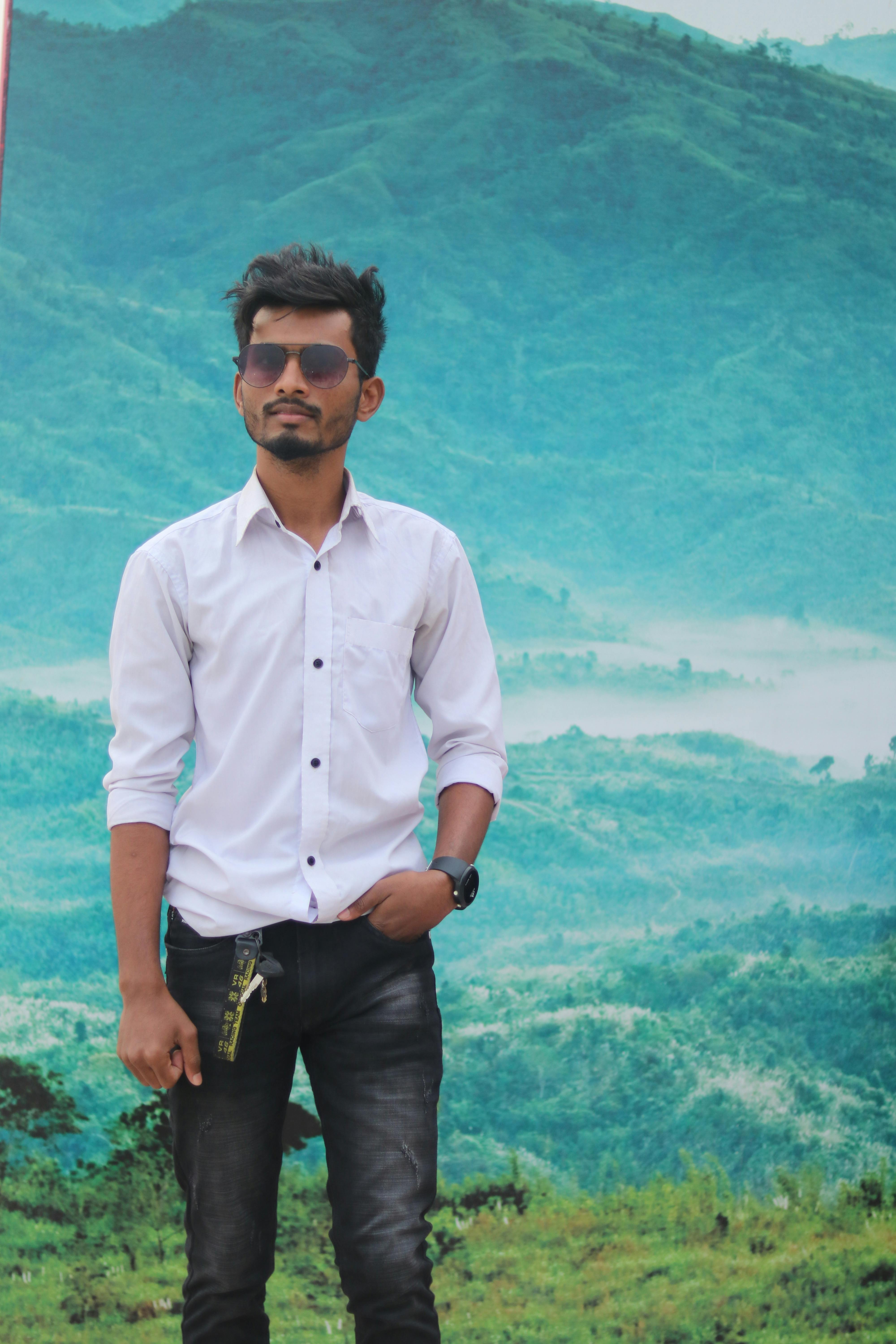 A stylish young man stands confidently against a scenic mountainous backdrop, wearing sunglasses and casual attire.