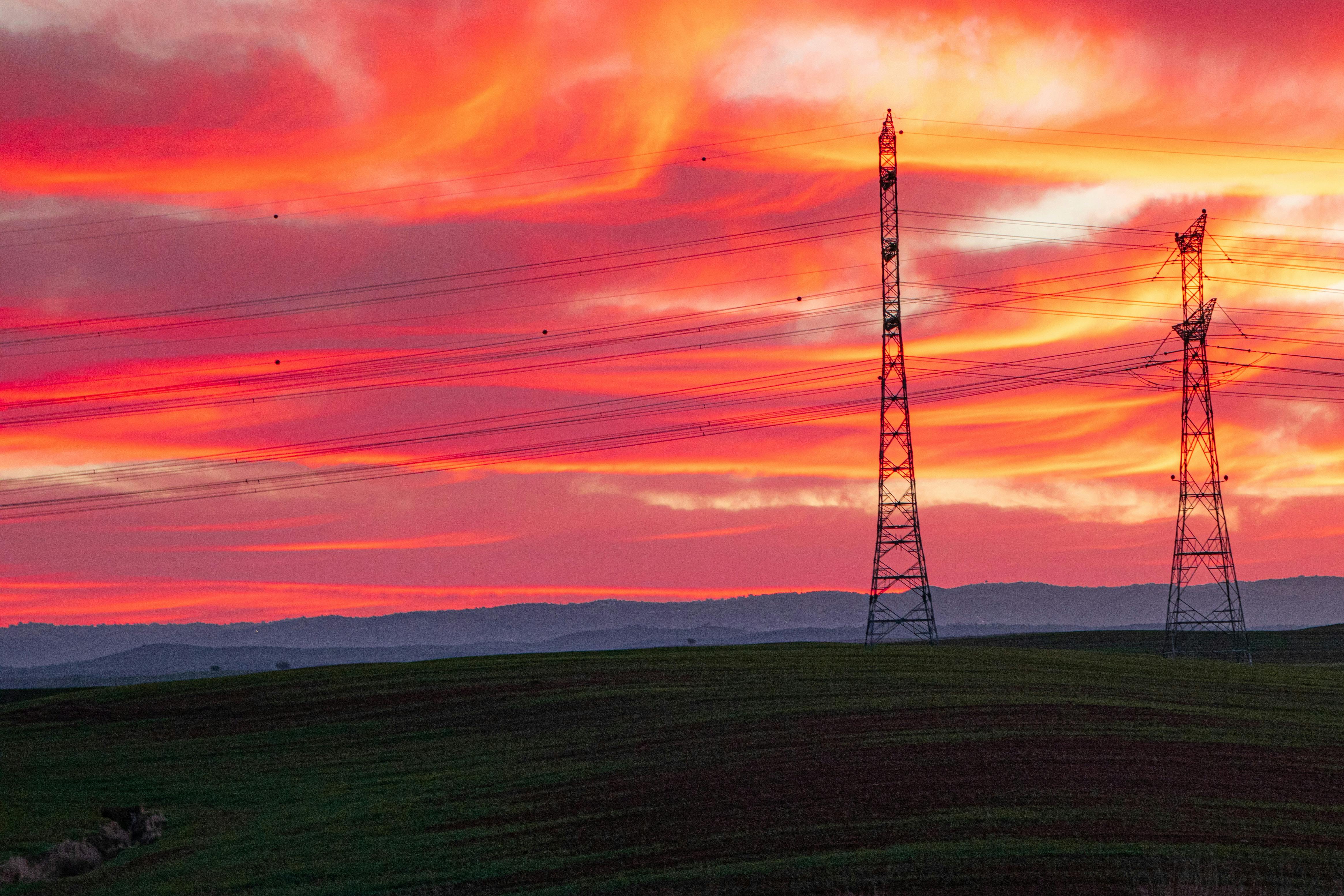 Electric pylons silhouetted against a colorful sunset sky over rolling hills.