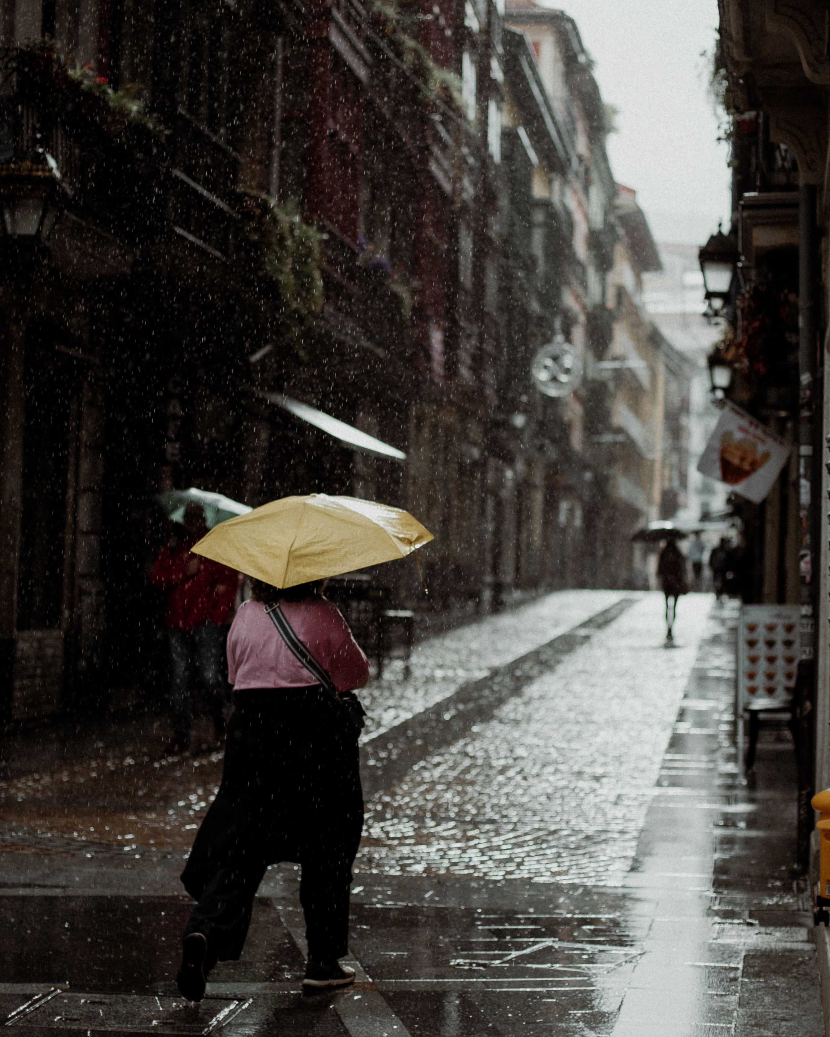 A person with a yellow umbrella walks down a cobblestone street in the rain, capturing a serene urban moment.