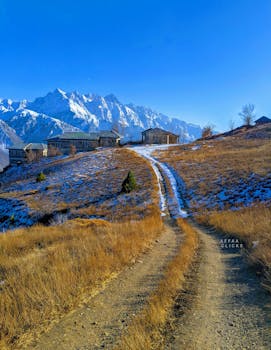 A dirt path leads to rustic houses with snowy mountains in Chitral, Pakistan.