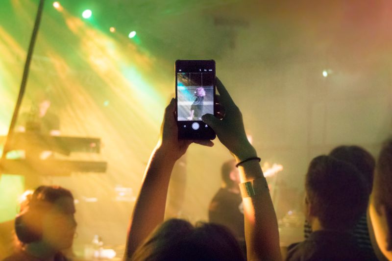 A lively concert crowd captures the performance with vibrant lights and a smartphone focus.