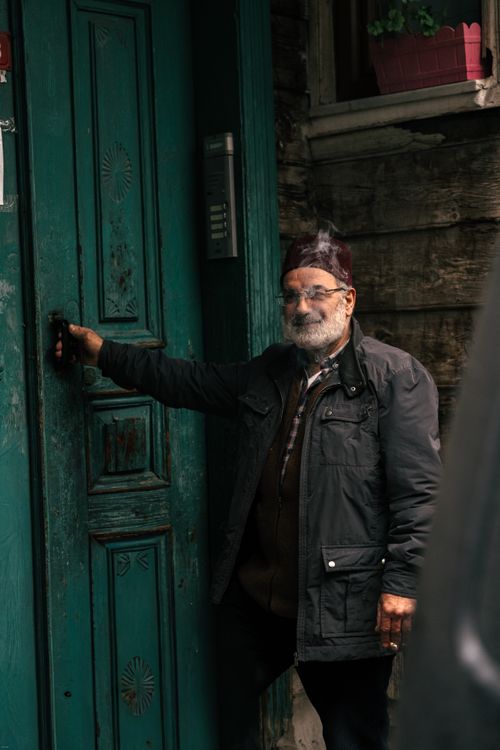 Free An elderly man with a hat stands smiling at a rustic green door, exuding a warm and welcoming vibe. Stock Photo