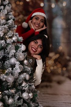 Two cheerful women celebrating Christmas by a decorated tree indoors.