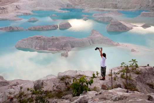 A woman takes a picture of a stunning blue lake amidst rocky terrain at sunrise.