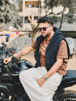 A fashionable young man wearing sunglasses sits on a motorcycle in an outdoor setting, exuding a cool and relaxed vibe.