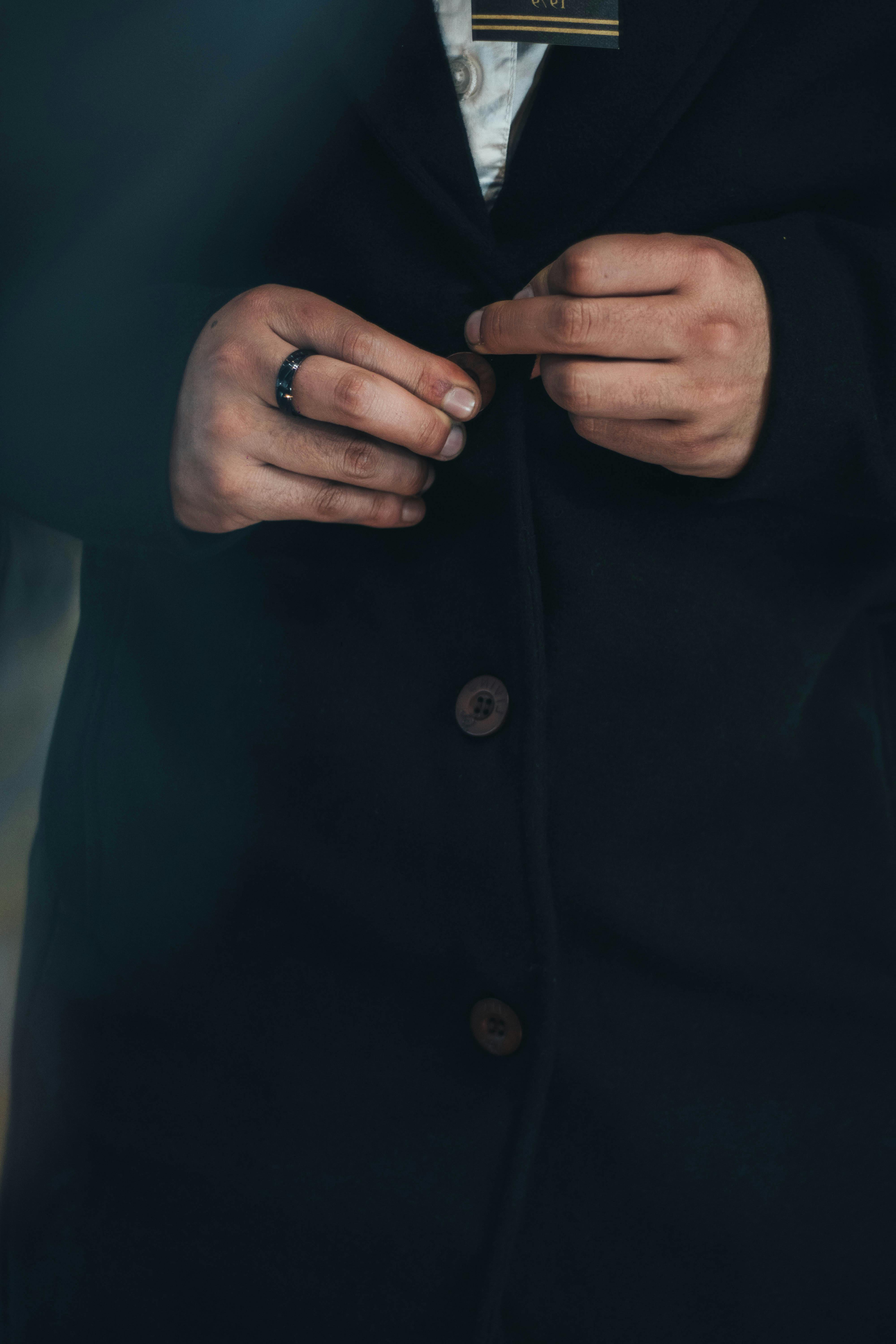 Gratis Un'immagine ravvicinata che ritrae delle mani che abbottonano un cappotto nero, in stile minimalista con un dettaglio ad anello. Foto a disposizione