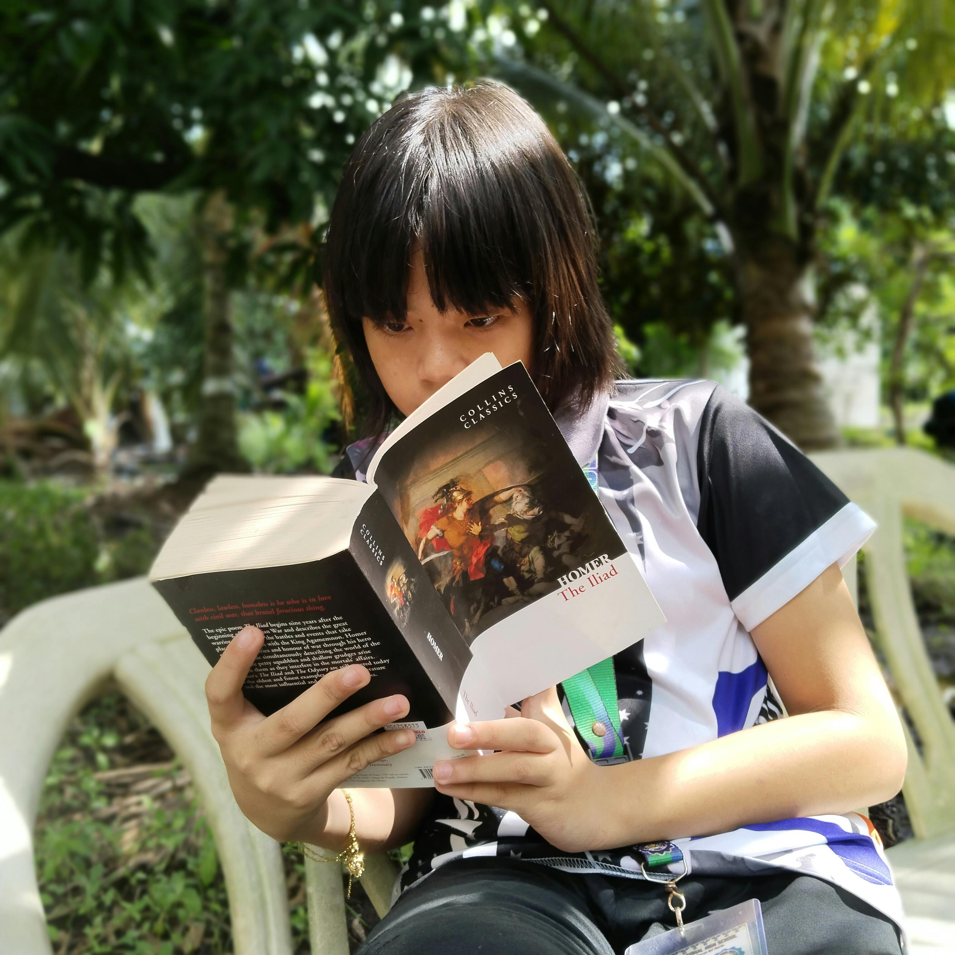 A teenager deeply engaged in reading 'The Iliad' by Homer outdoors in a lush setting.