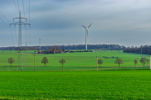 A tranquil landscape featuring green fields, a wind turbine, and power lines under an overcast sky.