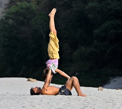 Two adults practicing acro yoga on a sandy beach with forest backdrop.